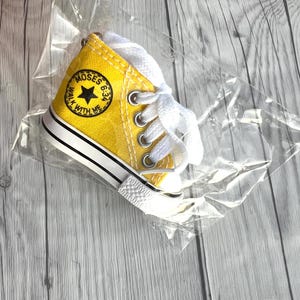 Puede incluir: Una zapatilla de lona amarilla en miniatura con cordones blancos, ribetes en blanco y negro y un parche redondo que dice "MOSES 6:34 WALK WITH ME". La zapatilla est&aacute; empaquetada en pl&aacute;stico transparente.