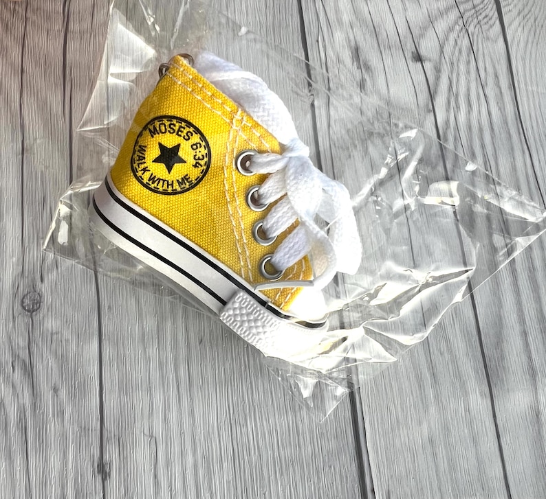 Puede incluir: Una zapatilla de lona amarilla en miniatura con cordones blancos, ribetes en blanco y negro y un parche redondo que dice "MOSES 6:34 WALK WITH ME". La zapatilla est&aacute; empaquetada en pl&aacute;stico transparente.