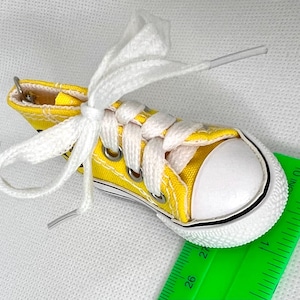 Puede incluir: Una zapatilla de lona amarilla en miniatura con cordones blancos y puntera blanca. El zapato peque&ntilde;o tiene ojales met&aacute;licos y una franja negra a lo largo de la suela. Una regla verde est&aacute; en primer plano.