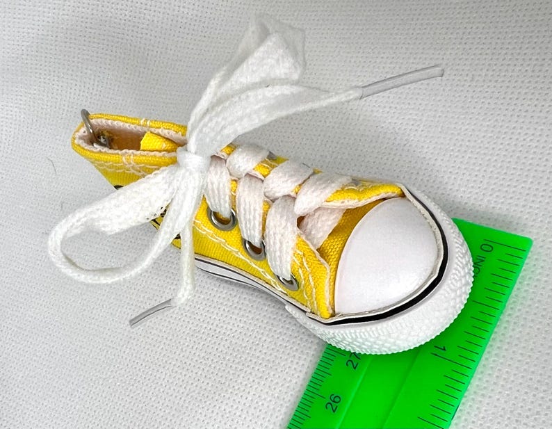 Puede incluir: Una zapatilla de lona amarilla en miniatura con cordones blancos y puntera blanca. El zapato peque&ntilde;o tiene ojales met&aacute;licos y una franja negra a lo largo de la suela. Una regla verde est&aacute; en primer plano.