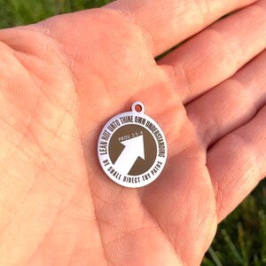 May include: A silver charm with a brown circle and a white arrow pointing upwards. The circle has the text "LEAN NOT UNTO THINE OWN UNDERSTANDING PROV 3.5.6 HE SHALL DIRECT THY PATHS".