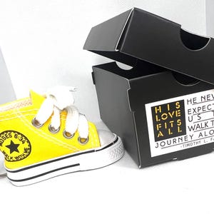 Puede incluir: Un llavero de zapatilla amarilla en miniatura con cordones blancos y suela negra y blanca. El llavero tiene el texto "MOSES 6:34 WALK WITH ME". Al lado hay una caja negra con el texto "HIS LOVE FITS ALL" y "HE NEVER EXPECTED US TO WALK THIS JOURNEY ALONE".