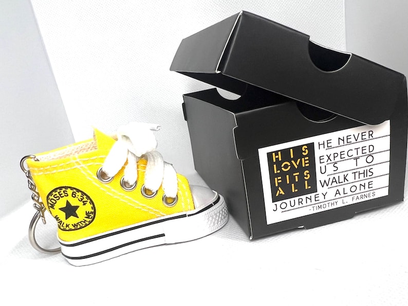 Puede incluir: Un llavero de zapatilla amarilla en miniatura con cordones blancos y suela negra y blanca. El llavero tiene el texto "MOSES 6:34 WALK WITH ME". Al lado hay una caja negra con el texto "HIS LOVE FITS ALL" y "HE NEVER EXPECTED US TO WALK THIS JOURNEY ALONE".