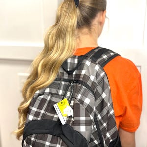 Puede incluir: Una mochila a cuadros negros, blancos y grises con detalles en negro. La mochila tiene una etiqueta adjunta. Una persona con cabello rubio largo en una cola de caballo lleva la mochila. Lleva una camiseta naranja.