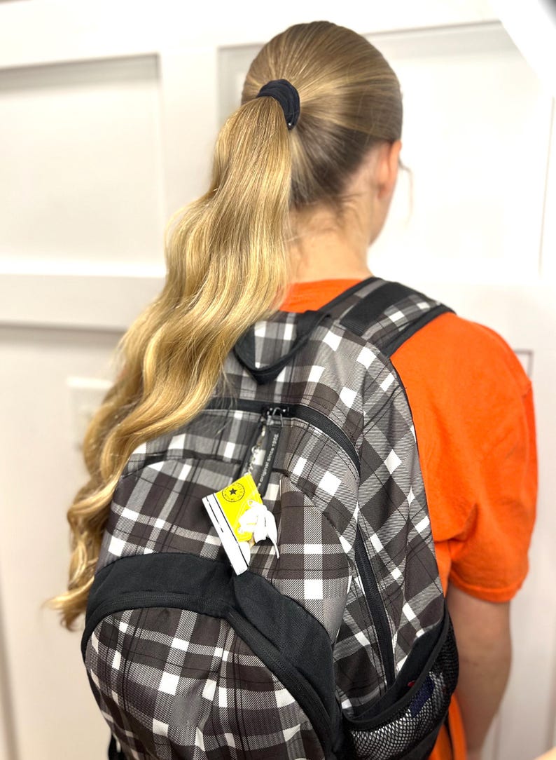 Puede incluir: Una mochila a cuadros negros, blancos y grises con detalles en negro. La mochila tiene una etiqueta adjunta. Una persona con cabello rubio largo en una cola de caballo lleva la mochila. Lleva una camiseta naranja.