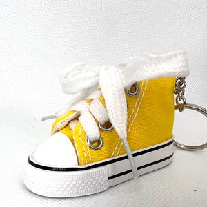 Puede incluir: Un llavero de zapatilla de lona amarilla en miniatura con cordones blancos y puntera blanca. El llavero tiene un anillo de metal plateado y una raya negra a lo largo de la suela. La zapatilla es de estilo bot&iacute;n.