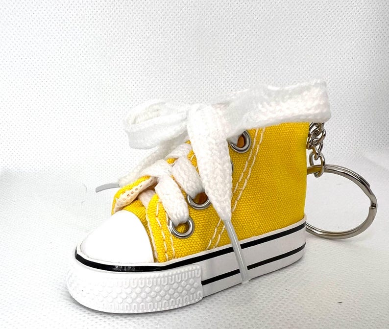 Puede incluir: Un llavero de zapatilla de lona amarilla en miniatura con cordones blancos y puntera blanca. El llavero tiene un anillo de metal plateado y una raya negra a lo largo de la suela. La zapatilla es de estilo bot&iacute;n.