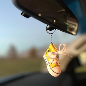 Puede incluir: Un llavero de zapatilla alta amarilla en miniatura con cordones blancos y suela blanca, colgado del espejo retrovisor de un coche. La zapatilla tiene un logotipo de estrella negra y un lazo de cinta blanca.