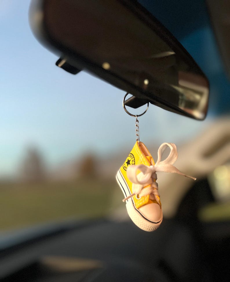 Puede incluir: Un llavero de zapatilla alta amarilla en miniatura con cordones blancos y suela blanca, colgado del espejo retrovisor de un coche. La zapatilla tiene un logotipo de estrella negra y un lazo de cinta blanca.