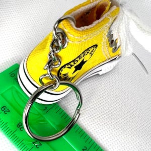 Puede incluir: Un llavero de zapatilla de lona amarilla en miniatura con suela negra y blanca, un llavero plateado y cordones blancos. La zapatilla tiene un gr&aacute;fico negro en el lateral. Una regla verde est&aacute; en primer plano.