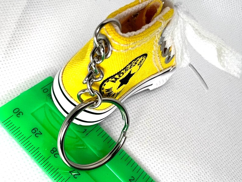 Puede incluir: Un llavero de zapatilla de lona amarilla en miniatura con suela negra y blanca, un llavero plateado y cordones blancos. La zapatilla tiene un gr&aacute;fico negro en el lateral. Una regla verde est&aacute; en primer plano.