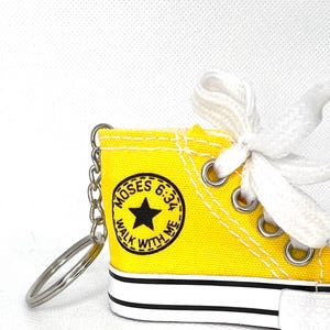 Puede incluir: Un llavero de zapatilla de lona amarilla en miniatura con cordones blancos y una cadena plateada. La zapatilla presenta una estrella negra y el texto "MOSES 6:34 WALK WITH ME" en un dise&ntilde;o circular. La suela es blanca con una franja negra.
