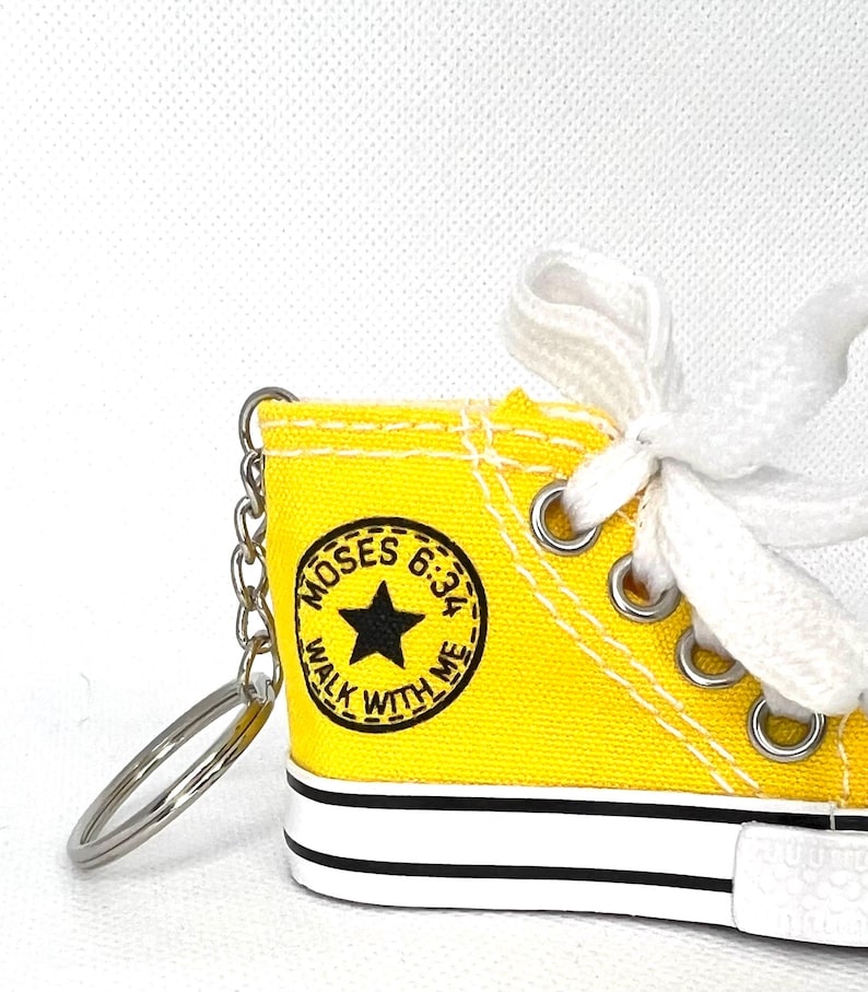 Puede incluir: Un llavero de zapatilla de lona amarilla en miniatura con cordones blancos y una cadena plateada. La zapatilla presenta una estrella negra y el texto "MOSES 6:34 WALK WITH ME" en un dise&ntilde;o circular. La suela es blanca con una franja negra.