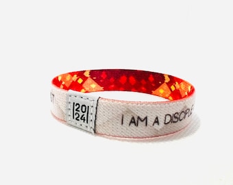 Könnte beinhalten: Weißes Armband mit rotem und orangefarbenem gemustertem Stoff auf der Rückseite. Das Armband trägt den Text "I AM A DISCIPLE" und die Zahlen "[20][24]" aufgedruckt.