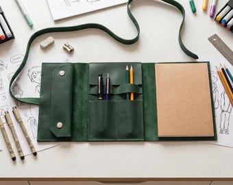 May include: A dark green leather roll-up organizer featuring a long strap, a flap with two silver snap buttons, multiple interior slots for pens and pencils, and a rectangular notebook with light brown paper pages.