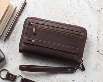 May include: A dark brown leather zip-around wallet with a wrist strap, a small notebook, a metal pen, black sunglasses, a pair of wireless earbuds in a charging case, and a leather keychain with metal keys.