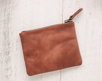 May include: A brown leather zippered pouch on a white wooden surface.