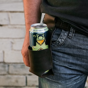 以下が含まれることがあります： 濃い茶色の革製ビール缶ホルダー。ベルトに取り付けられ、緑と青の缶が入っています。缶にはロゴとテキストがあります。人物はブルージーンズと黒いシャツを着ています。