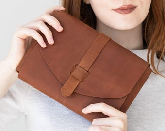 May include: A medium brown leather clutch bag featuring a smooth matte finish, a rounded flap closure, and a vertical leather strap detail that secures the front. The bag has a simple rectangular shape with clean lines and a minimalist aesthetic.