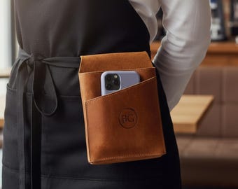 May include: A brown leather apron pocket with a phone inside. The pocket is attached to a black apron. The apron is worn by a person in a white shirt. The pocket has the initials "BG" embossed on it. The background is a cafe.