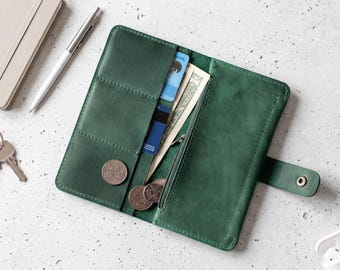 May include: An open forest green leather wallet featuring multiple card slots, a vertical pocket containing paper currency, and a zippered compartment. The wallet includes a snap button closure and contains several loose metallic coins.