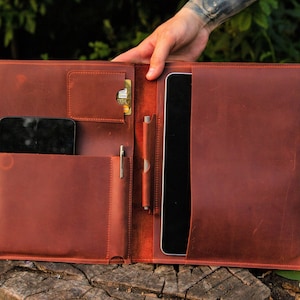 May include: A brown leather portfolio with a flap that opens to reveal a pocket for a tablet, a smaller pocket for a phone, and a pen holder.
