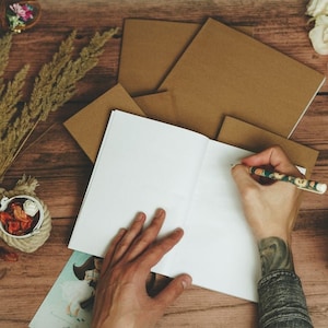 Puede incluir: Una persona escribiendo en un cuaderno en blanco con un l&aacute;piz marr&oacute;n sobre una mesa de madera. Hay sobres marrones, un ramo de hierba seca, una taza peque&ntilde;a de caf&eacute; y una botella de vidrio decorativa sobre la mesa.