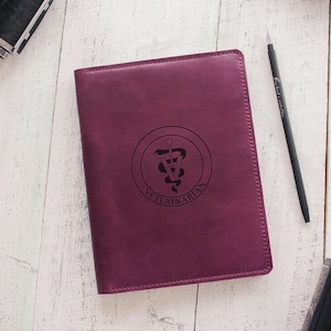 Può includere: Un diario in pelle viola con un logo in rilievo di un serpente avvolto attorno a un bastone con la parola "VETERINARIAN" sotto. Il diario è su una superficie di legno bianco.