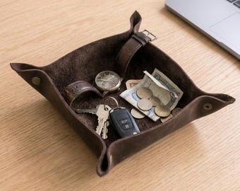 May include: A dark brown leather valet tray with snap-button corners containing a wristwatch with a brown leather strap, a set of metal keys, a black car key fob, several silver-colored coins, and a folded paper banknote.