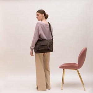 May include: A woman wearing a light purple sweater and beige pants is carrying a brown leather messenger bag with silver buckles. The bag is worn over her left shoulder. A pink chair with gold legs is in the background.