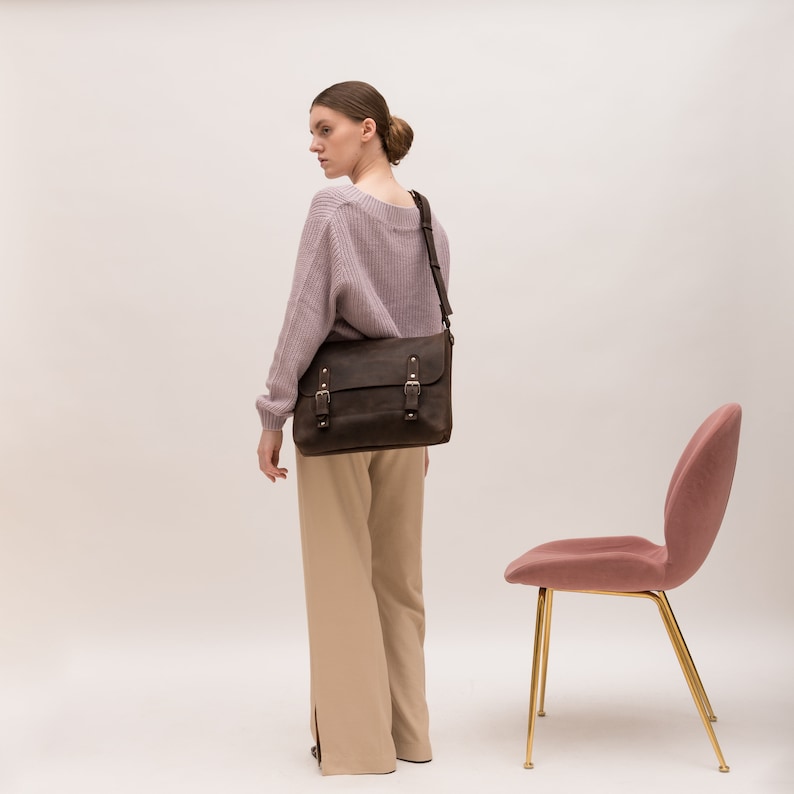 May include: A woman wearing a light purple sweater and beige pants is carrying a brown leather messenger bag with silver buckles. The bag is worn over her left shoulder. A pink chair with gold legs is in the background.