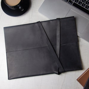 May include: A black leather laptop sleeve with a fold-over flap and a strap closure. The sleeve is lying flat on a white surface.