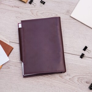 May include: A burgundy leather notebook cover with a white interior. The cover has a flap that folds over the front of the notebook.