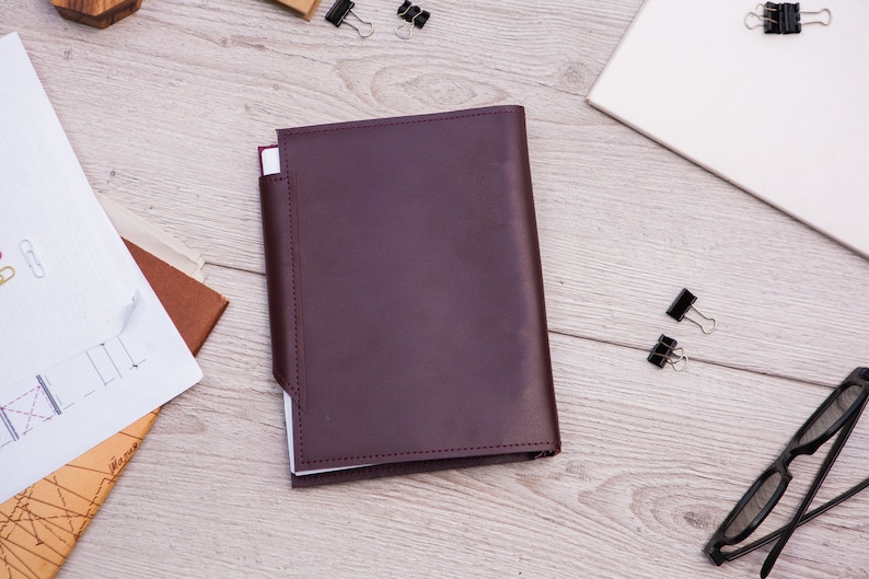 May include: A burgundy leather notebook cover with a white interior. The cover has a flap that folds over the front of the notebook.