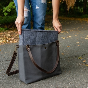May include: A grey canvas tote bag with brown leather straps and a grey felt lining. The bag is open and the lining is visible.