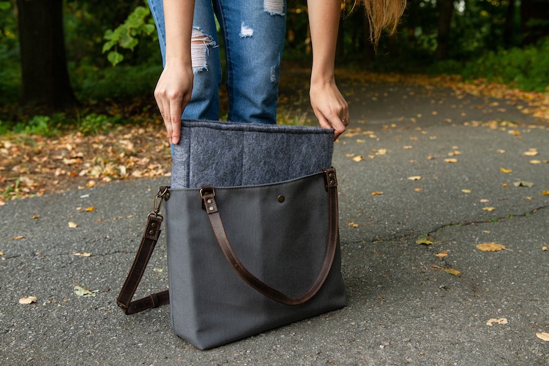 May include: A grey canvas tote bag with brown leather straps and a grey felt lining. The bag is open and the lining is visible.