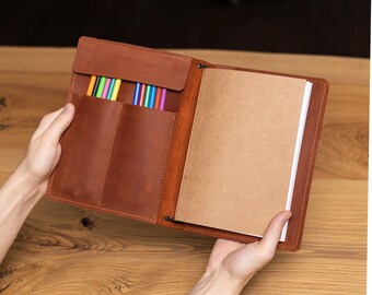 May include: Open brown leather notebook with a pen holder containing colorful pens. The notebook has a flap closure and a blank notepad. The notebook is held open, revealing the interior.