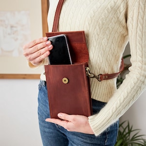 May include: A brown leather crossbody phone bag with a flap and a snap closure. The bag has a long, adjustable strap and a brass-colored clasp. A black smartphone is visible inside the open bag.
