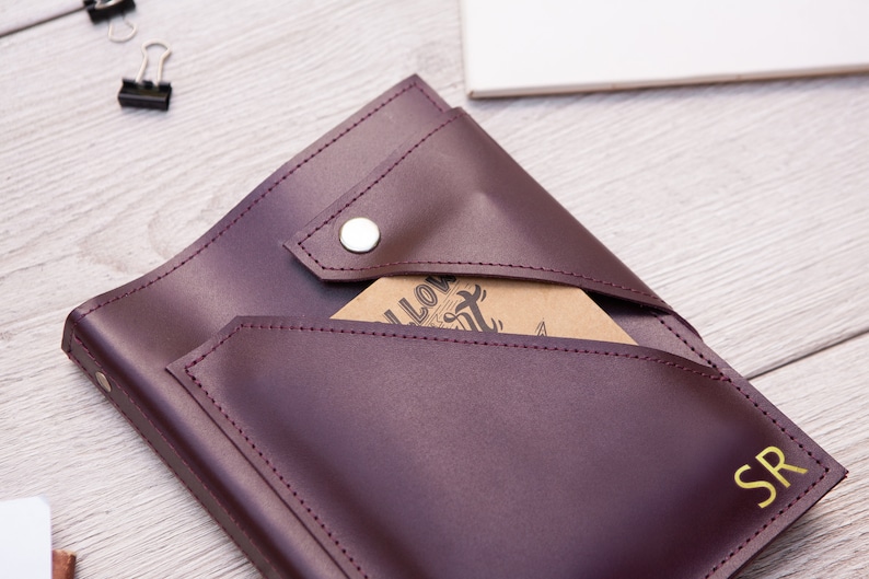 May include: Burgundy leather journal with a flap closure and a snap button. The journal has a pocket on the front and the initials "SR" are embossed on the front cover.