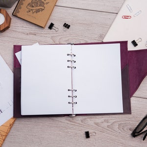 May include: A burgundy leather bound journal with a blank white page open. The journal has a silver ring binder and a flap closure. The journal is on a wooden surface with other office supplies.