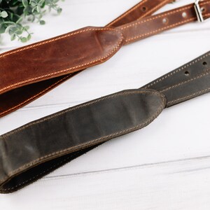 Peut inclure: Deux sangles de cam&eacute;ra en cuir, l'une marron et l'autre marron fonc&eacute;, avec des d&eacute;tails de couture. Les sangles sont dispos&eacute;es sur une surface en bois blanc.