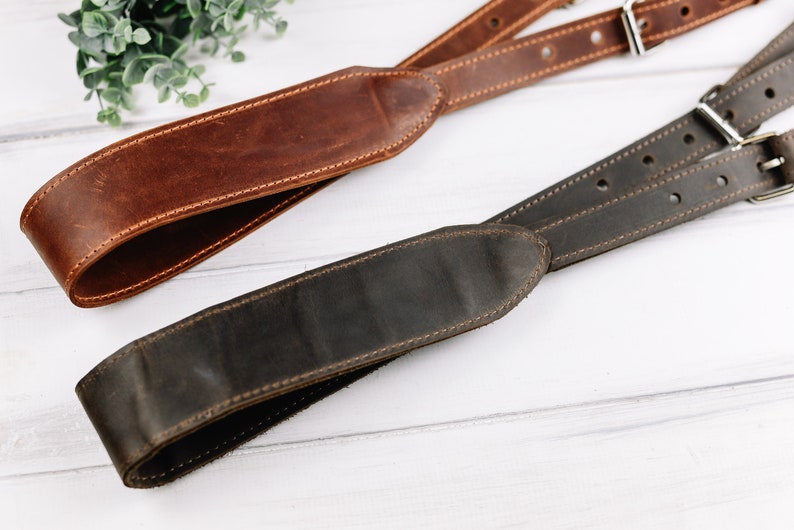 Peut inclure: Deux sangles de cam&eacute;ra en cuir, l'une marron et l'autre marron fonc&eacute;, avec des d&eacute;tails de couture. Les sangles sont dispos&eacute;es sur une surface en bois blanc.