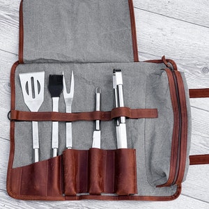 May include: A gray canvas and brown leather grilling tool roll with six tools, including a spatula, brush, fork, tongs, and two knives.