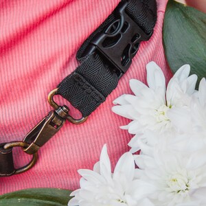 May include: A close-up of a black camera strap with a metal clasp. The strap is lying on a pink fabric surface with white flowers.