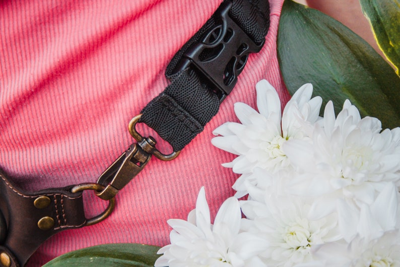 May include: A close-up of a black camera strap with a metal clasp. The strap is lying on a pink fabric surface with white flowers.