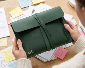 May include: A dark green leather document folder with a wrap-around strap closure. The front of the folder features the gold embossed initials MC in the bottom left corner. The folder has a smooth, matte texture and a rectangular shape.