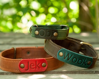 Puede incluir: Tres collares de perro de cuero con etiquetas de nombre personalizadas. Los collares son marrón, verde y azul verdoso. Las etiquetas de nombre son rojas, negras y azul verdoso. Los nombres en las etiquetas son "Jeko", "Obi" y "Wooko".