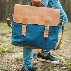 May include: A blue and brown messenger bag with a leather flap and accents. The bag has a canvas body with leather straps and buckles. It features a shoulder strap and a top handle. The bag is being carried outdoors.