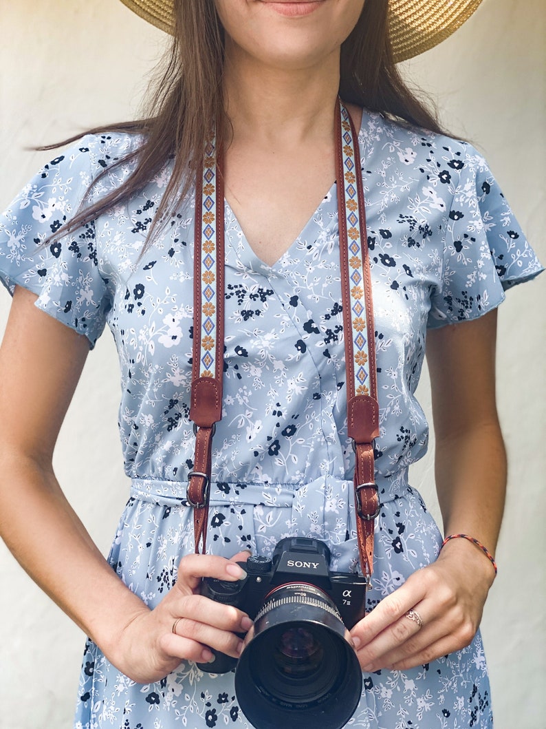 Puede incluir: Una mujer con un vestido azul floral y un sombrero de paja sostiene una c&aacute;mara Sony negra con una correa de c&aacute;mara marr&oacute;n y azul.