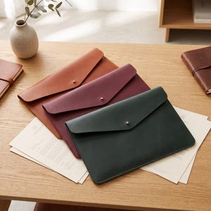 May include: Three flat document cases in tan, burgundy, and dark green leather, each featuring a pointed envelope-style flap secured with a single small gold-toned metal stud closure. The cases are arranged in a stack on top of white papers.